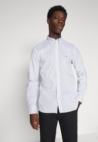 Camicia bianca con pois blu, maniche lunghe e colletto a punta; presenta un piccolo logo sul petto. Pantaloni neri.