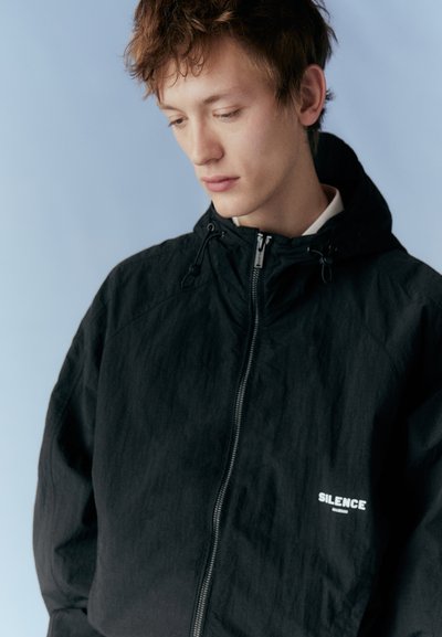 Black hooded jacket made of textured material, featuring a front zipper, adjustable drawstrings, and a printed logo reading "SILENCE."
