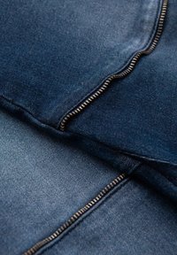 Tessuto di denim scuro con cuciture ben visibili e una cerniera evidente. La texture è liscia, con una leggera lucentezza in alcune aree.