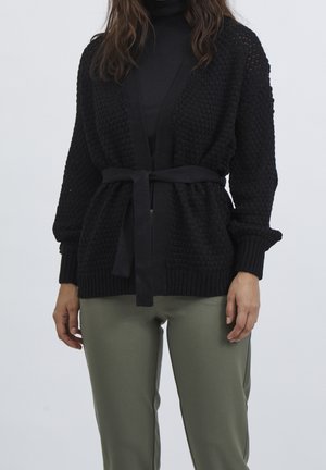 Femme portant un cardigan en tricot texturé noir avec une ceinture nouée à la taille, par-dessus un col roulé noir et un pantalon vert olive.