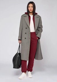 Grauer zweireihiger Mantel über weißem Oberteil und burgunderroten Jogginghosen. Das Modell hält eine schwarze Tasche und trägt weiße Sneaker.