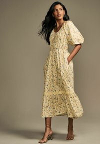 Abito midi floreale giallo chiaro con maniche a sbuffo, corpetto arricciato, tasche laterali e orlo svasato. Abbinato a tacchi beige con cinturini.
