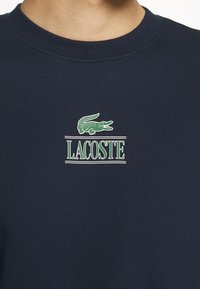 Sudadera azul marino con textura de canalé, que presenta un logo de cocodrilo verde y "LACOSTE" impreso en verde claro. Cuello de tripulación.