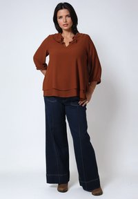 Vrouw met donker haar, gekleed in een roestkleurige blouse met een rimpelcol, brede donkere jeans en bruine laarzen, staand met haar handen op haar heupen.