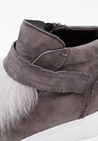 Bottine en suede grise avec une sangle texturée, ornée d'un accent en fausse fourrure gris clair et d'une semelle en caoutchouc blanc. Détail zippé à l'arrière.