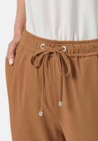 Bruine, zachte stoffen broek met een elastische tailleband, voorzien van een trekkoord met metalen aglets en zijzakken.