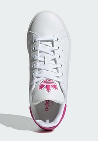 White leather sneaker with pink accents, round toe, perforated sides, and flat white laces. Features a logo on the tongue and heel.