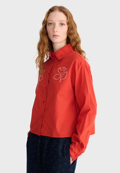 Jeune femme aux longs cheveux ondulés porte une chemise orange boutonnée avec une broderie de feuilles et un pantalon foncé à motifs, debout devant un fond uni.