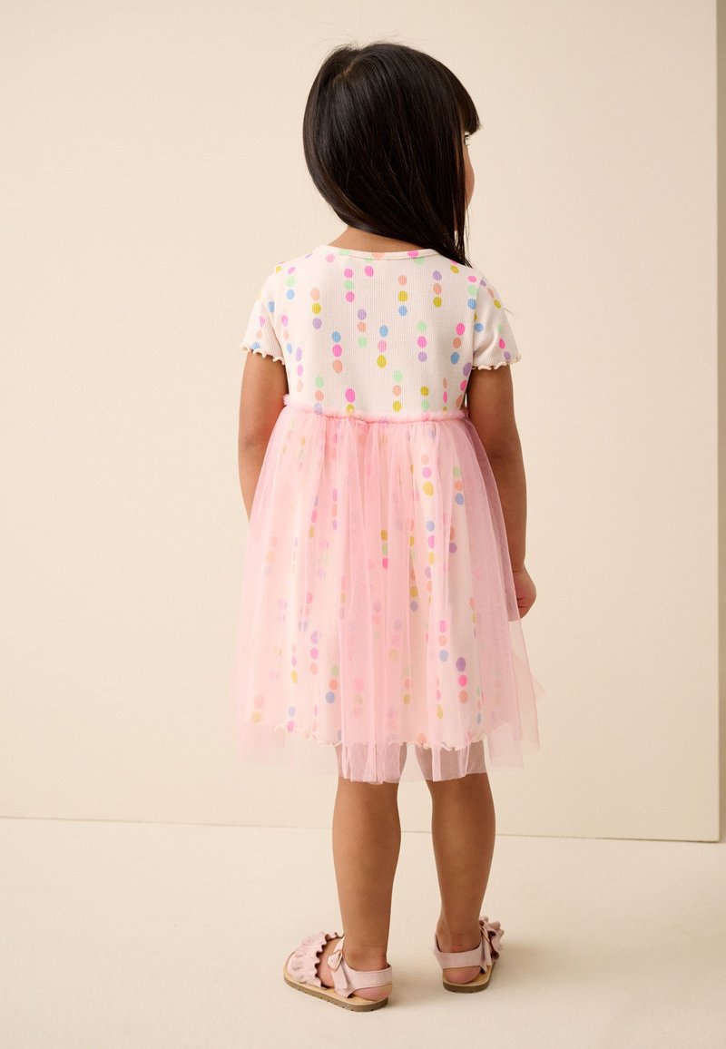 Niña joven con cabello oscuro que lleva un vestido blanco con puntos multicolores y una falda de tul rosa, de pie de espaldas sobre un fondo claro.