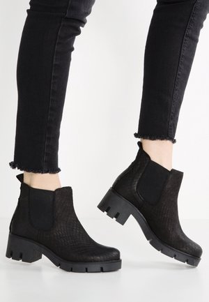 Personne portant des bottines noires texturées à talons épais et un jean noir ajusté avec des ourlets effilochés sur un fond blanc uni.