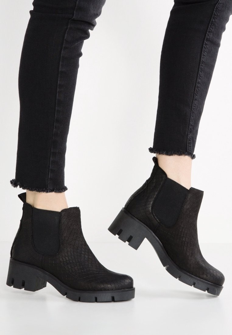 Personne portant des bottines noires texturées à talons épais et un jean noir ajusté avec des ourlets effilochés sur un fond blanc uni.