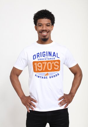 Homme souriant debout, les mains sur les hanches, portant un t-shirt blanc avec le texte bleu et orange « Original and Unrestored 1970's Vintage Goods ».