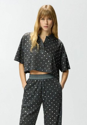 Donna che indossa una polo nera corta e pantaloni abbinati con pois argento, in piedi con le mani nelle tasche su sfondo semplice.