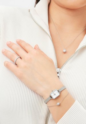 Srebrna mrežasta ura s kvadratnim številčnicom, v kombinaciji z nežno zapestnico iz verižice, ki vsebuje okroglo, teksturirano obesek. Roka počiva na svetli rebrasti tkanini.