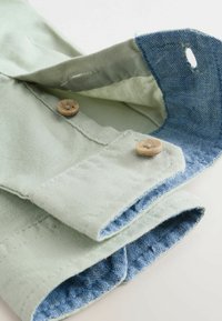 Manica in tessuto verde chiaro con bottoni in legno; il polsino interno presenta una texture in denim blu. Linee pulite e materiali a contrasto.