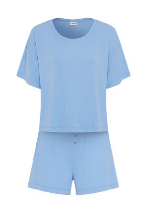Kvinde med langt mørkt hår, stående barfodet iført løs lyseblå kortærmet top og matchende shorts mod en ensfarvet lys baggrund.