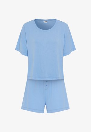 Kvinde med langt mørkt hår, stående barfodet iført løs lyseblå kortærmet top og matchende shorts mod en ensfarvet lys baggrund.
