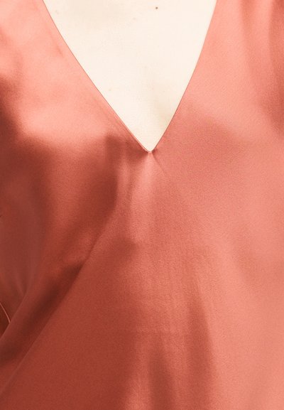 Μεταξένιο ένδυμα σε χρώμα σκουριάς με V-λαιμόκοψη σε φόντο ανοιχτού τόνου δέρματος, που δείχνει ομαλή υφή και ελαφρές πτυχώσεις του υφάσματος.