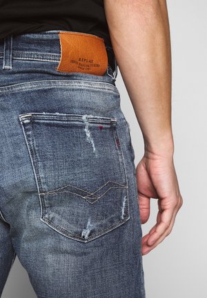 Blaue Denim-Jeans mit einem verwitterten Textur. Mit einem braunen Lederpatch mit der Aufschrift "REPLAY" und charakteristischer Naht an den Gesäßtaschen.