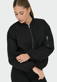 Veste bomber noire avec fermeture éclair à l'avant, col côtelé et poignets élastiqués. Poche supplémentaire sur la manche gauche. Texture lisse.