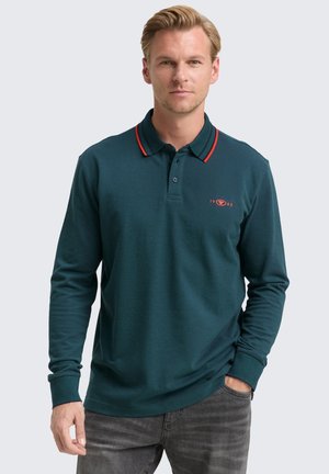 Man in een teal poloshirt met lange mouwen, met rode en marineblauwe kraagstrepen en geborduurde "1962" detail, gecombineerd met grijze jeans.