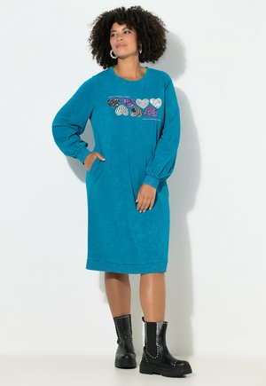 Mujer con cabello rizado vestida con un vestido de manga larga teal con patrones de corazones y botas de tobillo negras con tachuelas, de pie con una mano en el bolsillo.