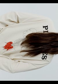 Krem košulja dugih rukava s crnim tekstom "PLEASE" djelomično vidljivim, crveni apstraktni cvjetni motiv i tamnosmeđa duga kosa koja prekriva dio leđa.