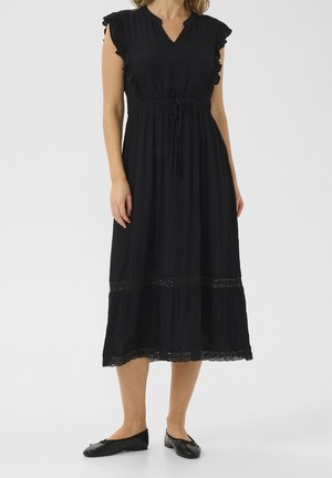 Femme portant une robe noire jusqu'aux genoux avec des manches courtes à volants, une taille ceinturée, une bordure en dentelle et des ballerines noires.
