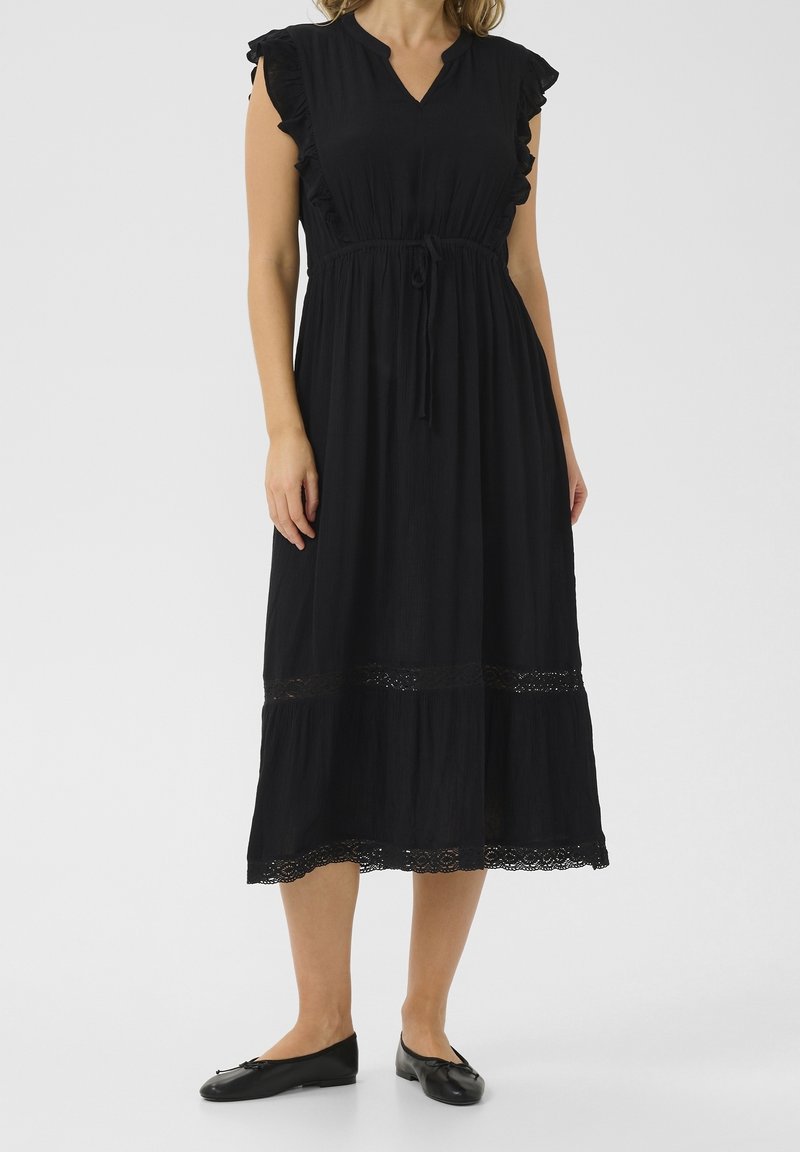 Femme portant une robe noire jusqu'aux genoux avec des manches courtes à volants, une taille ceinturée, une bordure en dentelle et des ballerines noires.