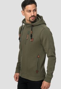 Sudadera con cremallera en color verde oliva, con un cuello alto, tejido texturizado, detalles en cuero, un bolsillo lateral con cremallera y detalles triangulares en los lados.