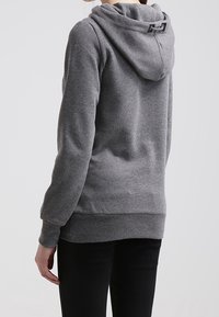 Harmaa huppari, jossa taitettu huppu, pehmeä pinta ja takaosassa oleva pieni merkki; yhdistetty mustien housujen kanssa. Normaali istuvuus, minimalistinen muotoilu.