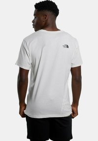 Camisa de algodón blanca con mangas cortas, que presenta un pequeño logotipo negro en la parte superior de la espalda y "The North Face" impreso en un tono más claro en la parte inferior.