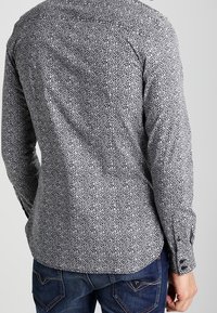 Chemise à manches longues à motif noir et blanc, coupe slim, avec un petit motif floral et des poignets boutonnés. Portée avec un jean bleu.