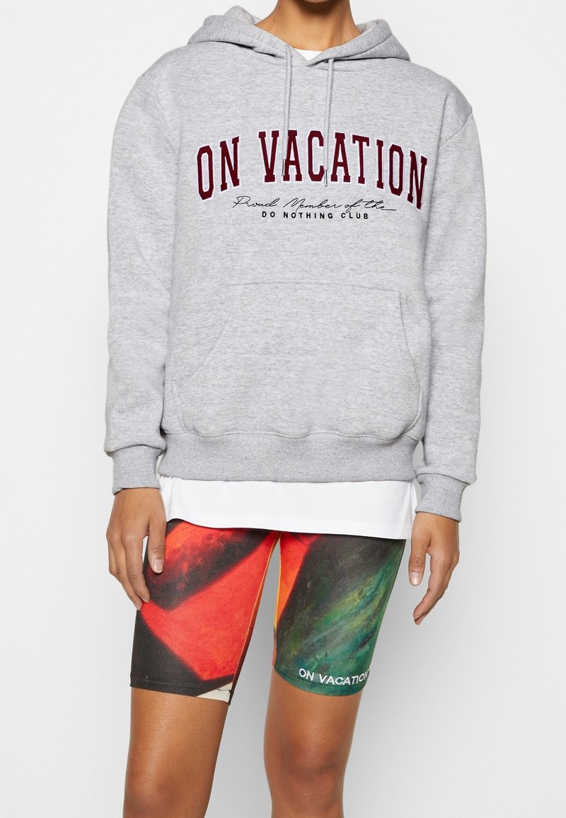 La personne porte un sweat à capuche gris avec le texte "ON VACATION" et un short multicolore avec le texte "ON VACATION" sur la cuisse gauche.