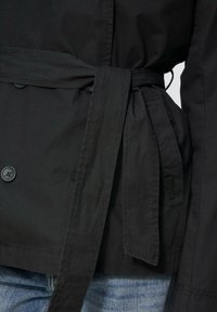 Schwarze Jacke mit Gurt, die eine glatte Textur aufweist und über Kn�öpfe, einen Bindegürtel und Seitentaschen verfügt. Sichtbare Nahtdetails am Saum.