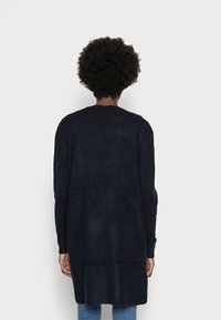 Pitkä, tummansininen kardigan, joka on valmistettu teksturoidusta neulemateriaaleista. Kardiganissa on rento istuvuus ja pidennetty helma, sekä rosoiset olkapäät.