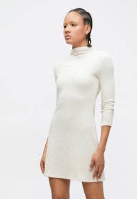 Rochie cu guler înalt, crem, cu dungi, mâneci trei sferturi, corsaj strâns și tiv evazat, realizată dintr-un material moale tricotat.