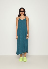 Robe maxi teal avec un motif floral, bretelles réglables, et une silhouette fluide, associée à des sabots verts vifs.