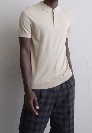 Mann trägt ein kurzärmeliges beiges Poloshirt und blau-braune karierte Hose und steht vor einer einfarbigen Wand.