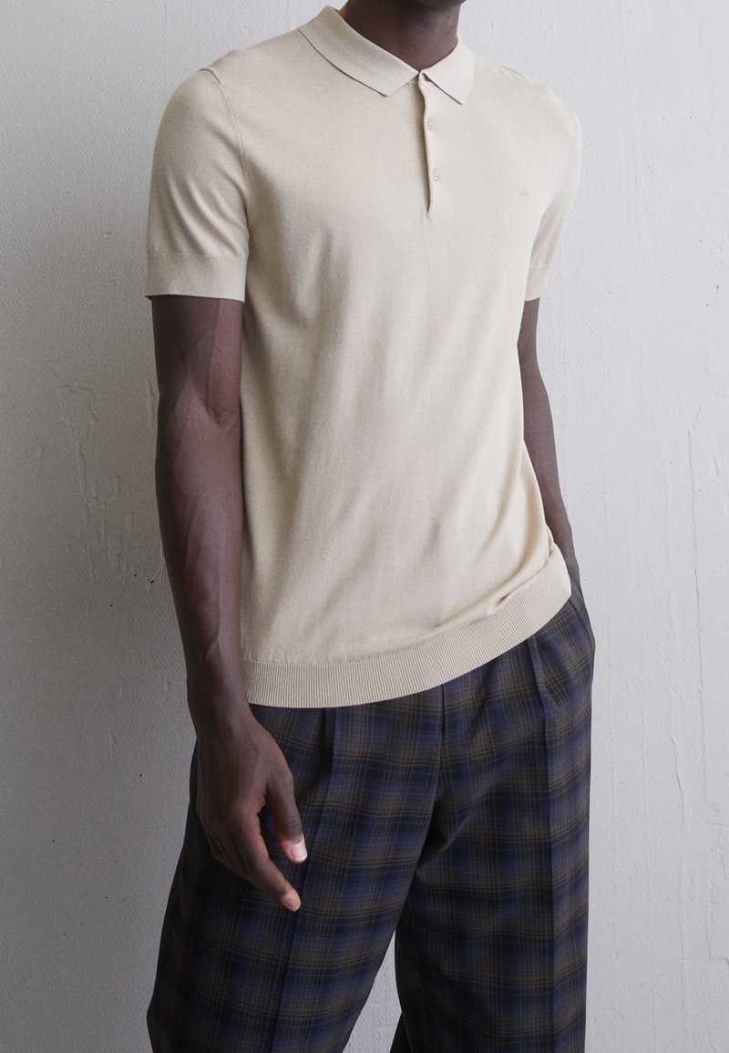 Homme portant un polo beige à manches courtes et un pantalon à carreaux bleu et marron, debout contre un mur uni.