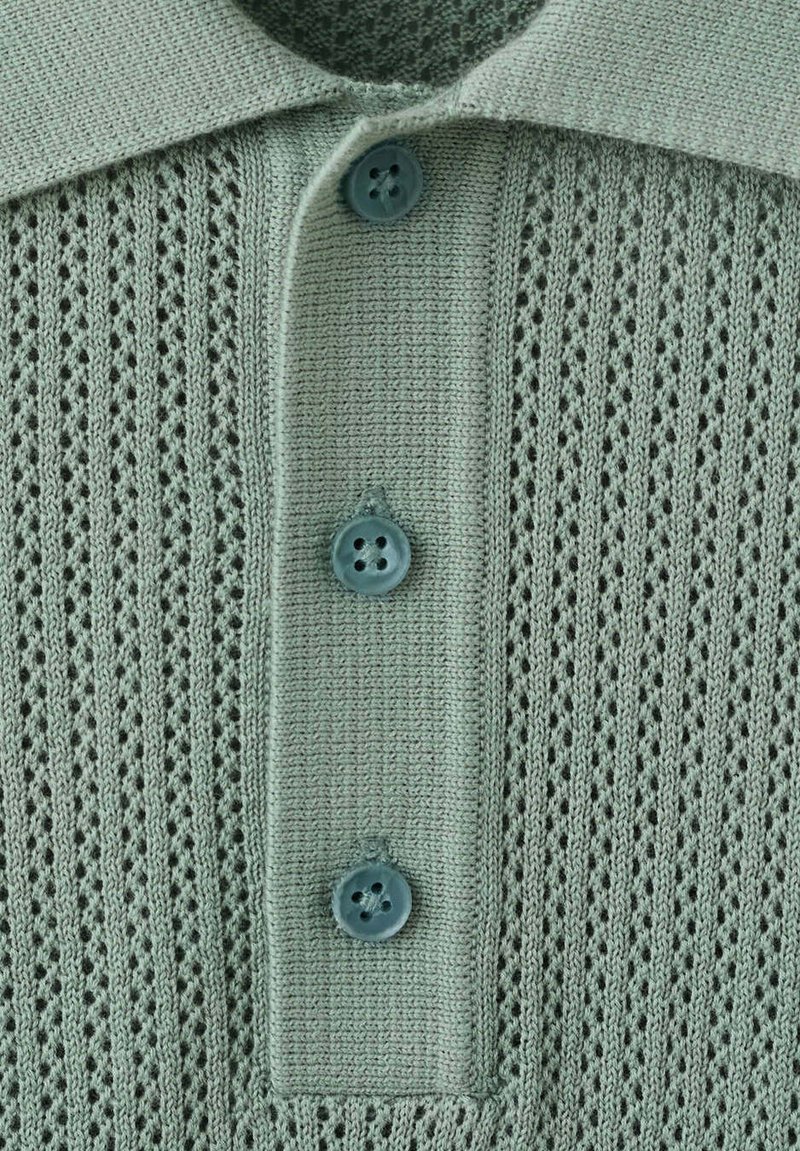 Chemise tricotée bleu clair avec un col et trois boutons bleus sur un tissu texturé à motif.