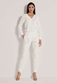 Witte lange mouwen v-hals top gecombineerd met hoge witte broek met plooien en enkelopeningen, gedragen met sandaaltjes met hakken.