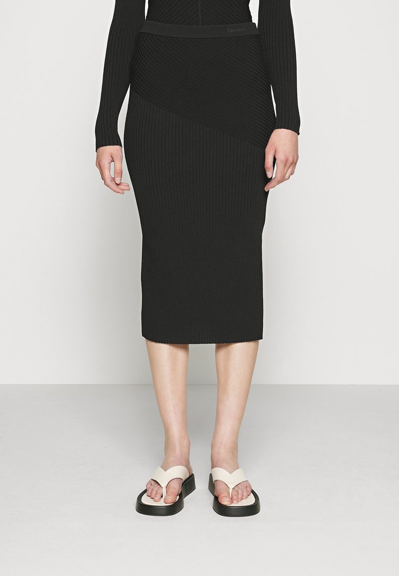 Jupe midi noire à côtes avec un design asymétrique, associée à des tongs crèmes avec une semelle noire épaisse. Texture lisse et coupe ajustée.