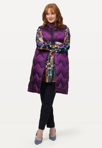 Lila Steppweste mit Zickzackmuster und hohem Kragen. Kombiniert mit einem mehrfarbigen gemusterten Hemd und dunklen Hosen, abgerundet mit blauen High Heels.