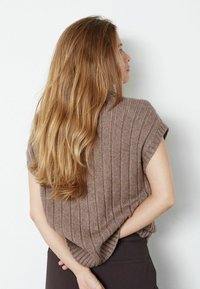 Hnědý pletený svetr s krátkým rukávem, s volným střihem a zaobleným dolním okrajem. Modelka je postavena s rukama sepjatýma za zády.