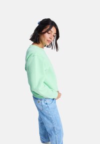 Sudadera verde menta con puños y dobladillo acanalados, que presenta un ajuste relajado. Combinada con jeans de corte holgado en azul claro. Textura suave.