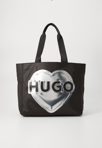 Melnā auduma soma, kas izgatavota no sintētiskā materiāla, ar lielu sudraba sirds dizainu un treknos burtos rakstītu vārdu "HUGO". Izturīgas melnas rokas.