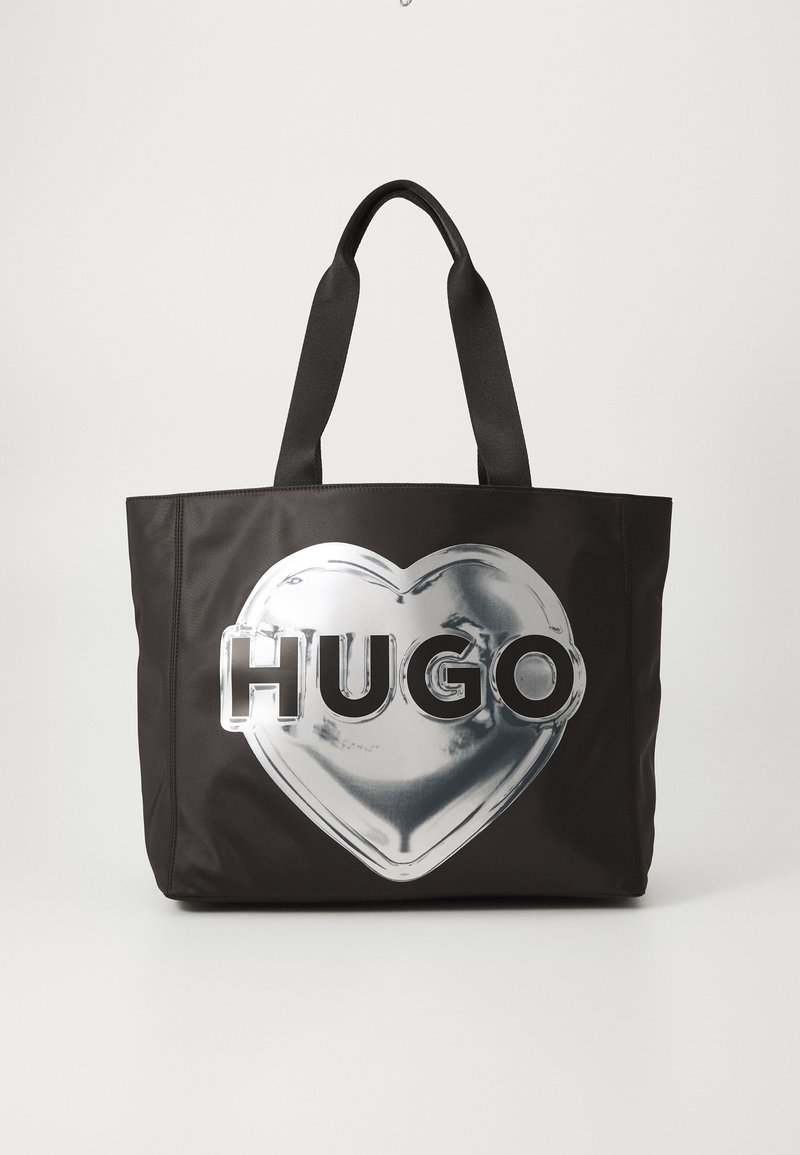 Melnā auduma soma, kas izgatavota no sintētiskā materiāla, ar lielu sudraba sirds dizainu un treknos burtos rakstītu vārdu "HUGO". Izturīgas melnas rokas.