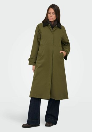 Femme portant un long manteau vert olive avec un col noir, un jean large foncé et des chaussures marron, posant avec une main dans la poche du manteau.