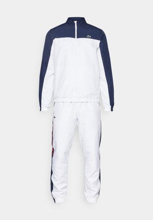 Tuta bianca con dettagli blu navy, composta da una giacca con zip e pantaloni con vita elasticizzata. Logo Lacoste sul petto e sulla gamba. Texture del tessuto liscia.